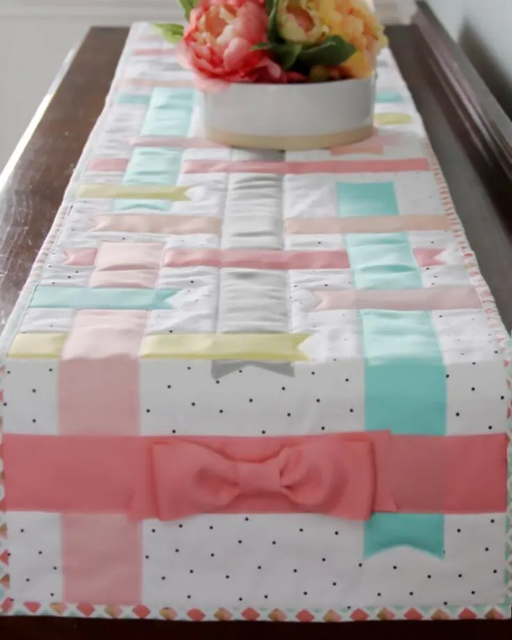 Don't you love how soft and calming this quilt runner is? It's a sweet and feminine design adorned with pastel ribbons and is excellent for lightening up the room for spring. It's a simple project that is great for beginners and involves straight-line sewing. Table runner size: Adjustable